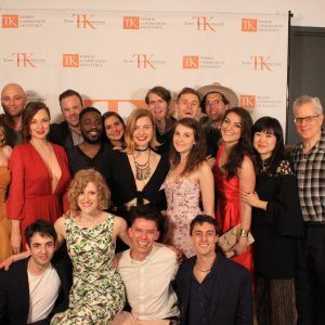 A group of Meisner Graduates is smiling and posing together at an event. They stand in front of a backdrop featuring "TK" logos and words, indicating a celebratory or formal gathering. Some are dressed in evening attire.