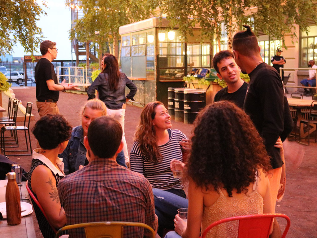 A group of people sits at an outdoor table, socializing and enjoying drinks on a warmly lit patio in NYC. With greenery and string lights enhancing the background, the lively atmosphere is perfect for those looking for fun summer things to do. Some stand and chat, fully immersed in this urban oasis.