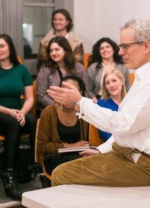 A man in a white shirt, seated on a desk, is speaking to a group of attentive people in an Open Studios classroom setting. The attendees are smiling and appear engaged, sitting on vibrant orange chairs. The scene conveys a sense of lively discussion and collaborative learning.