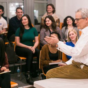 A man in a white shirt, seated on a desk, is speaking to a group of attentive people in an Open Studios classroom setting. The attendees are smiling and appear engaged, sitting on vibrant orange chairs. The scene conveys a sense of lively discussion and collaborative learning.