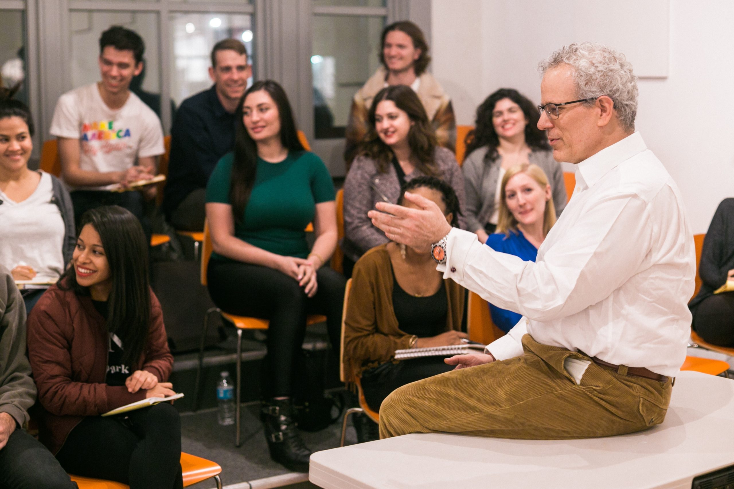 A gray-haired man in a white shirt sits on a table, gesturing animatedly like William Esper, as he talks to an engaged group of people around him. The group, consisting of both men and women, takes notes in the brightly lit room.