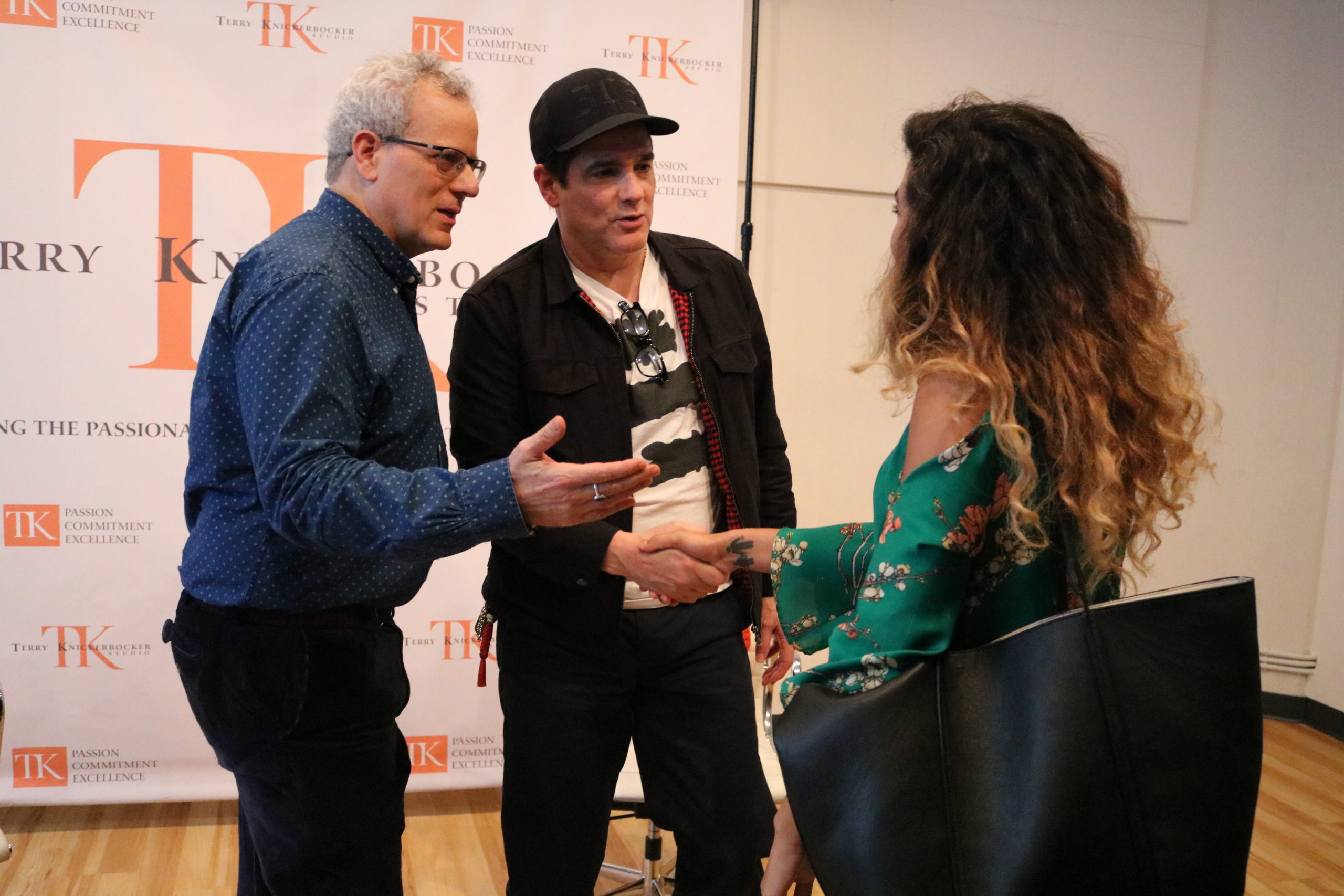 Three people are engaging in conversation. Two men, one resembling Yul Vazquez of Russian Doll fame, stand on the left as a woman in a green floral dress stands on the right. They are inside a room with a "TK" branded backdrop, likely linked to Terry Knickerbocker Studio. The men gesticulate animatedly as they talk.