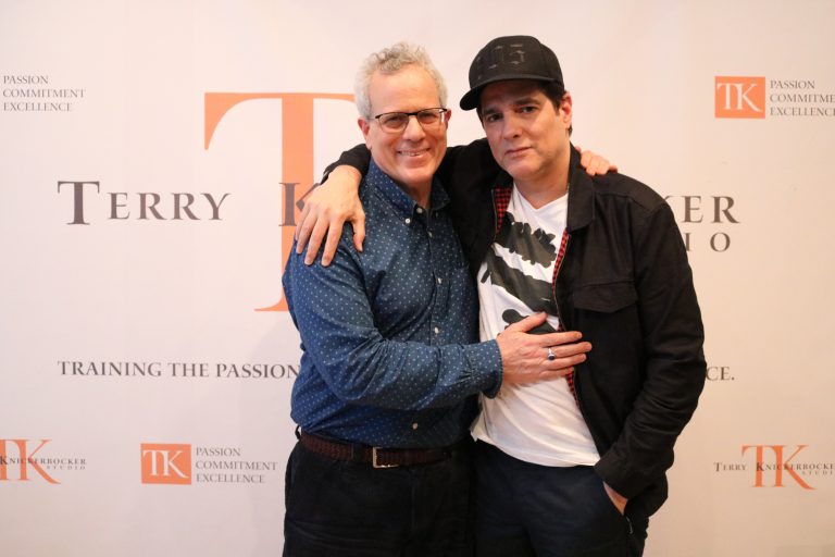 Two people stand together in front of a backdrop that reads "Terry Knickerbocker Studio" with "Passion, Commitment, Excellence." One wears a cap and graphic t-shirt, the other dons glasses and a button-up shirt. Could it be Yul Vazquez from Russian Doll showcasing his versatile style?