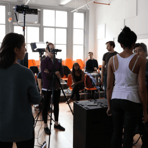 A group of people is gathered in a well-lit room, some seated on orange chairs and others standing. A camera setup is visible, capturing the focused individuals immersed in an engaging session of on-camera acting.