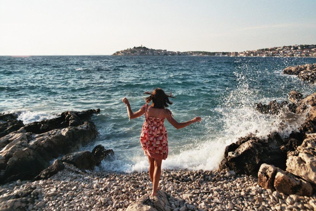 A woman in a red patterned dress stands on the authentic rocky shores, facing the ocean with a town in the background. Waves crash against the rocks under a clear sky.