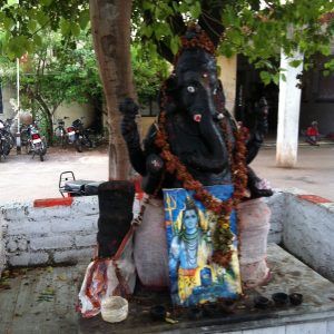 A small outdoor shrine, a traveler's serene discovery, features a statue of Ganesha adorned with garlands under a tree. At its base lies a framed image of Lord Shiva. In the background, motorcycles hint at the journey's continuation.