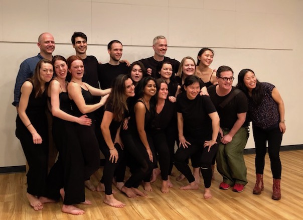 A group of sixteen people, wearing casual black attire, are gathered indoors on a wooden floor. They are smiling and posing together for a group photo. Some are standing while others kneel in front, embodying the relaxed and friendly atmosphere of their creative process at work.