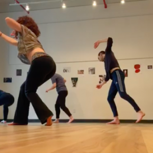 In a vibrant community dance class set in a studio with wooden floors, participants are barefoot, each striking a dynamic pose. The walls showcase various small artworks and photographs, creating an inspiring atmosphere akin to actor training sessions.