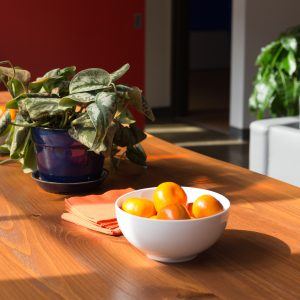 A white bowl filled with bright orange fruits sits on a wooden table beside folded orange napkins, creating a harmonious feng shui balance. A potted plant with green leaves graces the background, where sunlight streams in, casting soothing shadows on the scene.