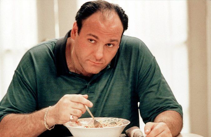 A person in a green shirt sits at a table, holding a spoon over a bowl of pasta, channeling their inner James Gandolfini, and looking directly at the camera. The setting suggests a casual dining environment with soft lighting.