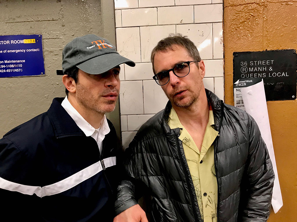 Two men in casual attire, perhaps actors known for frequent collaborations, stand against a tiled wall in a subway station. One wears a cap and jacket, while the other sports sunglasses and a puffer jacket. A sign indicating "36 Street M & N & Q trains" is partially visible on the wall.