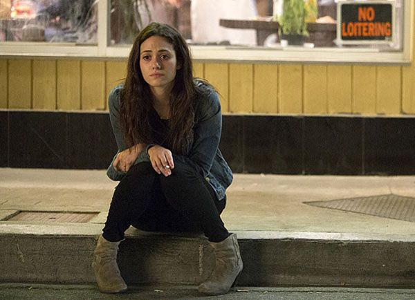 A woman with long hair sits on a curb, embodying particularization in her denim jacket, black pants, and light-colored boots. A "No Loitering" sign hangs noticeably in the window behind her as she appears thoughtful or concerned.