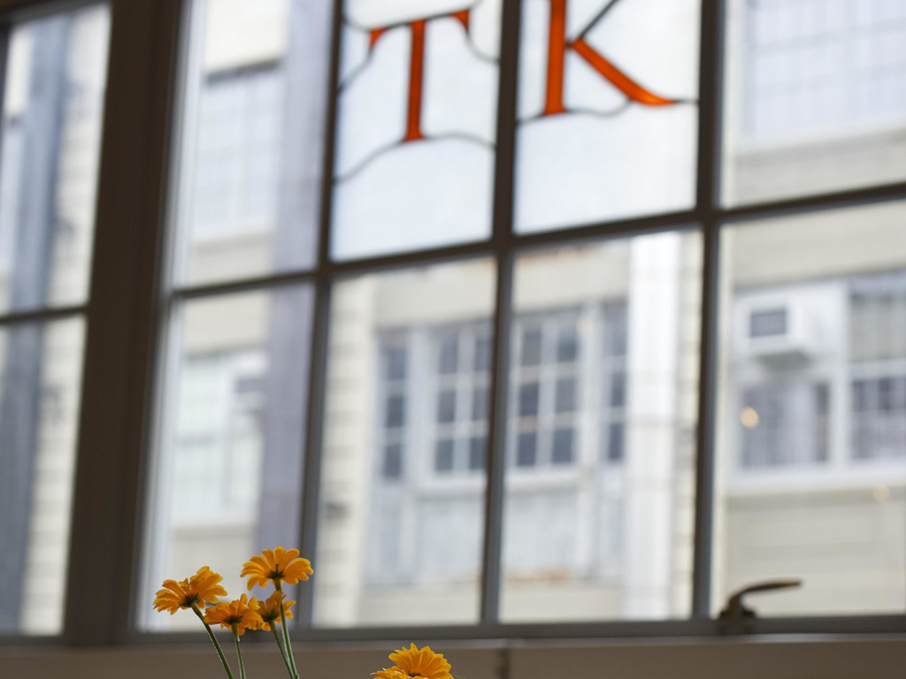 Small yellow flowers in a vase sit on a windowsill, exuding an optimistic charm. A large window behind them reveals a blurred view of urban buildings, while the letters "T" and "K" form part of a creative stained glass design at the top of the window.