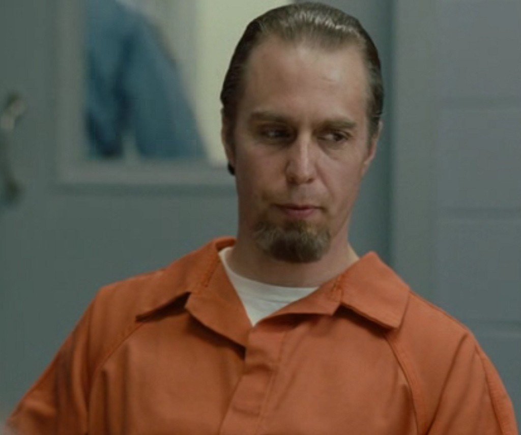 A man resembling Sam Rockwell, with a goatee and wearing an orange prison jumpsuit, sits in a room with a serious expression. The background is blurred, showcasing a blue and gray color scheme.