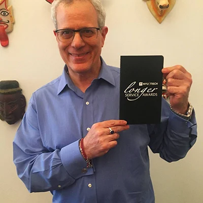 A person in a blue shirt proudly displays a black book emblazoned with "NYU Service Award" from the prestigious Tisch Longevity Program. They stand before a wall adorned with decorative masks, adding to the celebratory atmosphere.