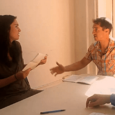 A man and a woman are sitting at a table in discussion with their acting coach. The woman is holding a piece of paper, while the man gestures with his hand. A pen and open notebook are also on the table. Bright sunlight filters into the room.