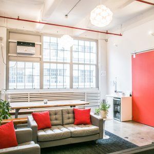 A modern living room with an authentic touch, featuring a grey couch adorned with vibrant red throw pillows. The space boasts large windows, an authentic red accent wall, and exquisite hanging light fixtures. A long table and potted plants complement the air conditioner on the wall.