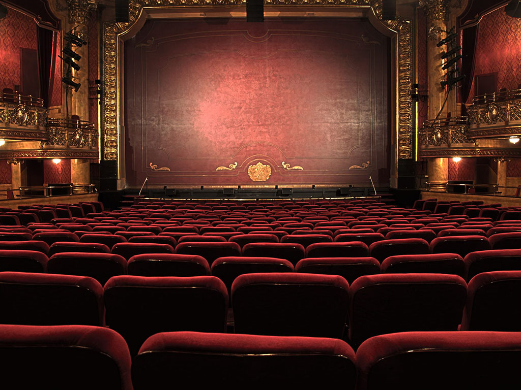 The affordable theater boasts rows of plush red seats facing a grand stage, framed by ornate gold detailing and a large, closed red curtain. Despite its budget-friendly prices, the interior impresses with intricate architectural elements and subtle lighting.