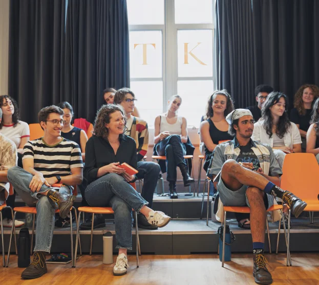 A group of actors sitting in a bright studio classroom, engaging and smiling during a workshop discussion, representing creative wellness in a supportive training environment.