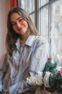 Stella Jiler leaning against a snowflake-decorated window with holiday greenery, wearing a striped shirt, featured as a Fall 2025 Student of the Season at Terry Knickerbocker Studio NYC.